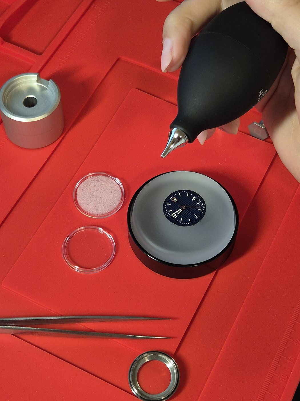 A watchmaker's hand using a bulb-style dust blower over a movement seated in a casing cup on a red benchmat, surrounded by tweezers and a case ring