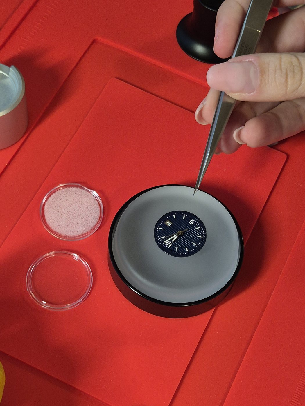 Watchmaker's tweezers positioned over an NH05 movement on a red benchmat, hour and minute hands installed on a navy blue wave dial