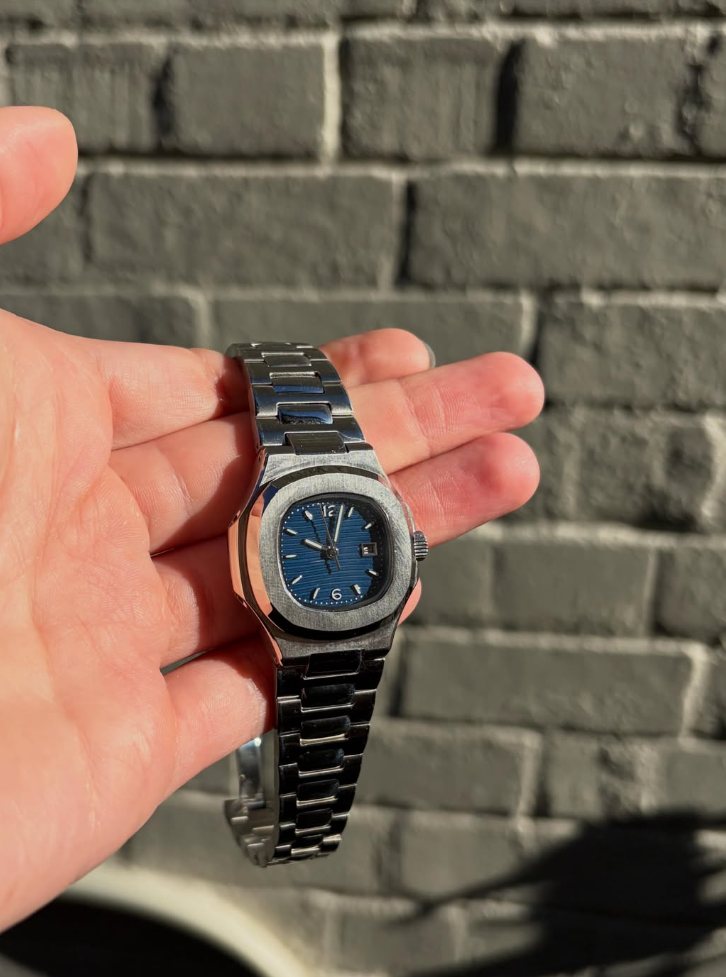A finished NH05 build in a Nautilus-style case with a navy blue wave dial and silver hands, held in a palm against a brick wall
